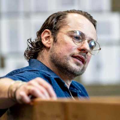 A close up shot of a person wearing glasses with mid-length hair in blue shirt sitting on the bench in a rehearsal room facing sideway 