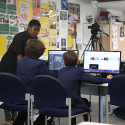 Two students sit at desks working at computers while an artist looks over the desks helping them. 