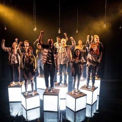 A group of people with angry expressions and their fists raised in the air stand on cubes emitting white light on a stage under six unlit hanging lightbulbs. 
