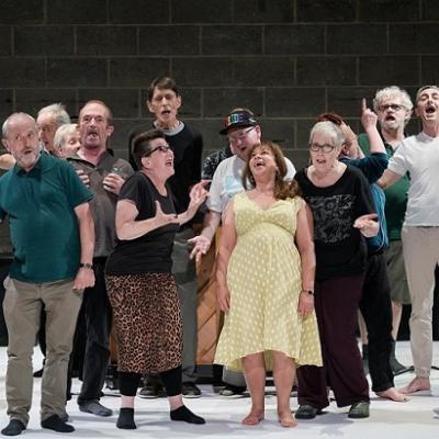 A large group of people standing on a white floor in front of a brick background, their mouths open as if in song.