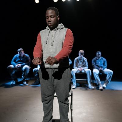 A young person in a hoodie and joggers ands in front of a chair with arms out, while behind them in the background five people in chairs are visible