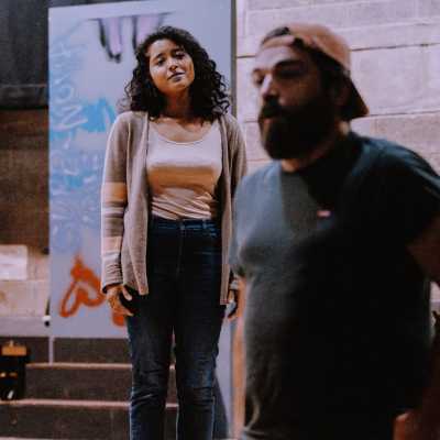 Two people in a rehearsal room. One stands with their back tot he other, and the one behind looks at him. 