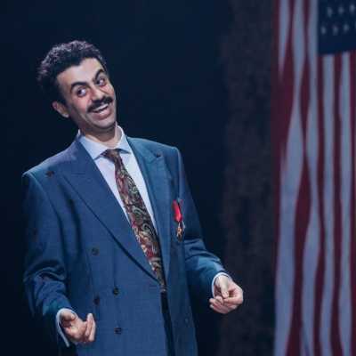 A man in a smart suit is smiling and looking to the side. He looks as though he has just made a joke. An American flag hangs behind him to one side. 