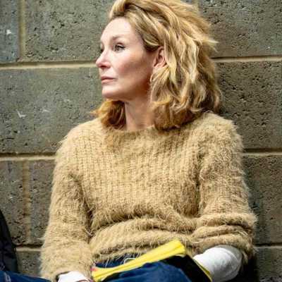 A close up shot of a white person with blonde long hair, wearing beige jumper and a pair of jeans sitting on the floor against the wall with legs crossed looking sideway 