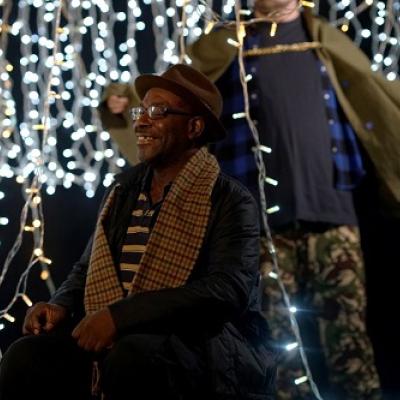 A person in a plaid scarf, black coat and brown hat stands smiling in a black space amongst hanging white fairy lights, while two other figures are visble blurred in the background.
