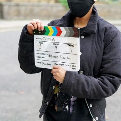 A person in a dark coat and black face mask standing outside holding a film clapperboard