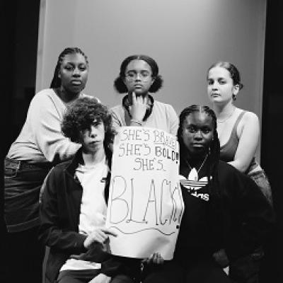 Five young people sit and stand with serious expressions in front of a white background, holding a sign that reads "She's brave! She's bold! She's Black!"