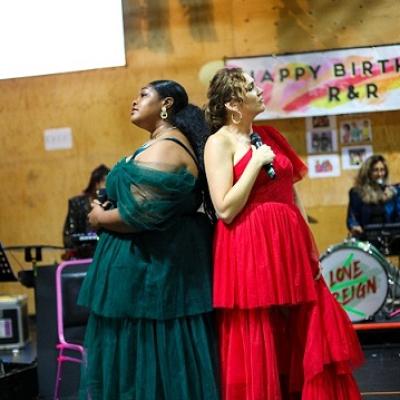 Two people in flowing green and red dresses stand back to back on a stage holding microphones with a band visible in the background.