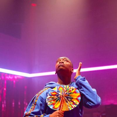 A person in a blue jumper holding a round brightly coloured fan looking up with an arm by their face in a room with a pink LED light strip around the walls and a pink fringe curtain