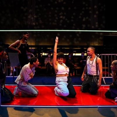 A group of people in ballroom outfits pose on a red runway with audience behind them