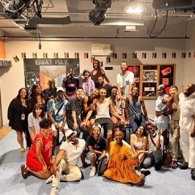 A large group of people pose for a picture in a studio space with pride flag bunting hanging around the walls 