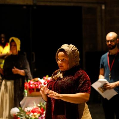 A person in a red embroidered cardigan and brown bonnet stands on a stage speaking, while in the background 3 other people are visible 