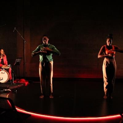 Two people stand on a round platform lit from below with red fluorescent light with their arms in front of their chests. On the left hand side, a band with a drummer is visible. 