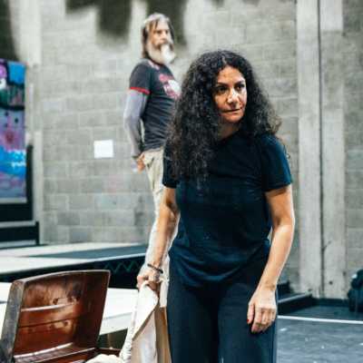 A person with long, dark hair stands in a theatre rehearsal room holding a burlap sack. 