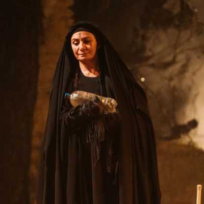 A woman dressed entirely in black with a black headscarf, holds a sand covered bottle of water in both hands. Behind her is smoke. 