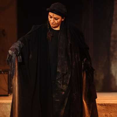 A woman dressed entirely in black with a black headscarf holds up one arm. Sand is pouring out of her robes onto the floor.