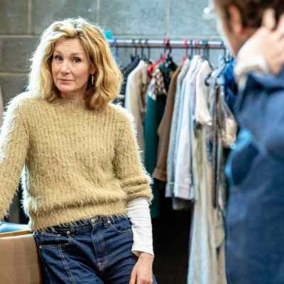 An actor with blonde mid-length hair standing leaning against a bench looking at the actor who's back to the camera in blue shirt 