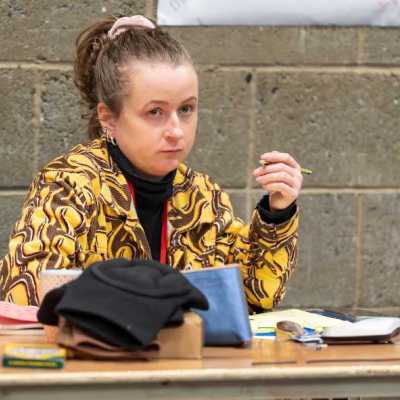 A young person sits at a table in a theatre rehearsal room, taking notes in a notebook