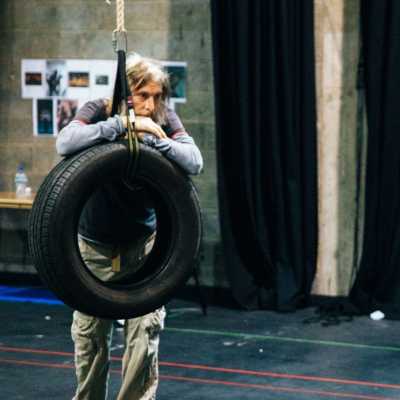 A person with a long beard wearing layered shirts leans against a tire swing in a theatre rehearsal room. 