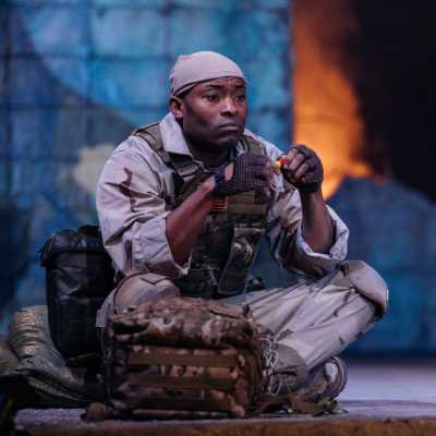 A soldier sits cross legged with his camouflaged bag in front of him and sand bags stacked to the side. He looks sad and thoughtful. 