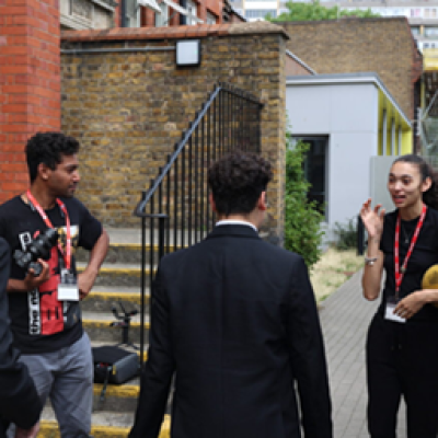 Two people, one holding a camera and one holding a football, speak to a student with their back to the camera. 