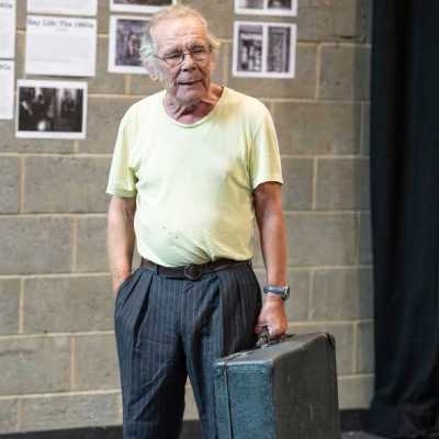 An older man holds an old-fashioned suitcase, standing in a theatre rehearsal room and performing a scene