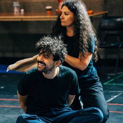 A person wearing jeans and a short-sleeved black shirt sits on the theatre rehearsal room floor with their hands behind their back. Another person sits behind them, holding a burlap sack. 