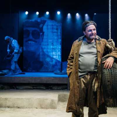 A man in a large overcoat leans with one arm wrapped around a car tyre that is suspended from the ceiling by rope. He looks off to the left with a slight smile on his face. Behind him in a blue light, an army soldier can be seen sorting his kit. 