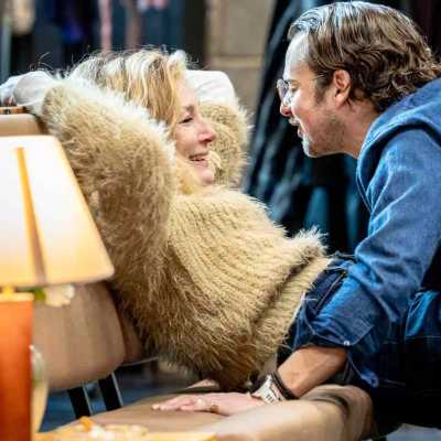 An actor with blonde mid-length hair standing leaning against a bench looking at the actor who's back to the camera in blue shirt 