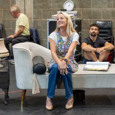 A blonde woman wearing a colourful apron sits on a white chaise couch in a theatre rehearsal room, performing a scene.  10 - A young man with short, curly dark hair, wearing a long-sleeved black shirt, and a blonde woman wearing a colorful apron, both sit on a white chaise couch in a theatre rehearsal room. They perform a scene together, holding scripts