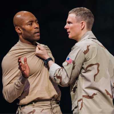 Two soldiers against a black backdrop. One has the other by the collar of his top, the other holds one hand up in protest.