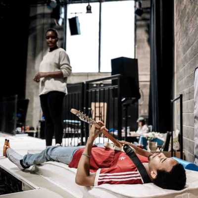 A slim, Afro-Caribbean man of average height with an Afro dressed in light blue jeans, white converses and a red t-shirt lies on a mattress holding a guitar. Behind him, an Black woman of average height with short hair wearing black leggings and a white jumper stands looking at him. Both actors are on a raised white latform with a black railing behind them, and a rehearsal room with a lighting rig on the ceiling, black speakers and large windows is visible in the background.