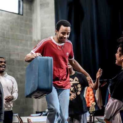 A slim, Afro-Caribbean man of average height with an Afro dressed in light blue jeans, white converses and a red t-shirt holding a blue suitcase walks across a line of chairs. Around him three other actors are partially visible singing towards him. In the background, a rehearsal room with concrete walls, long black curtains and a window is visible.