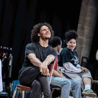 A man of average height with light brown skin and a big afro wearing a black t-shirt and dark gray joggers sits on a chair in a rehearsal room with his legs crossed and his his hands clasped on his knees, smiling. Around him four more people stand and sit, some of whom are holding scripts, and behind him a rehearsal room with concrete walls, long black curtains and a costume rack is visible.