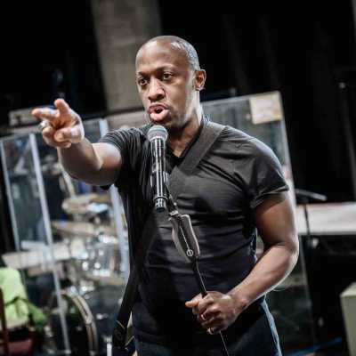 A Black man of average height wearing a black t-shirt with a guitar slung behind his back on a strap stands singing into a microphone. He holds the mic stand at an angle in one hand, and the other arm is extended forward with index and middle fingers out. Behind him a rehearsal room with concrete walls, black curtains and various music equipment like a drumkit, guitars and mic stands are visible. 