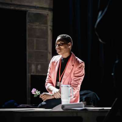 A Black woman with short hair sits in a room with a dark background. She wears a pink jacket and holds a single flower.