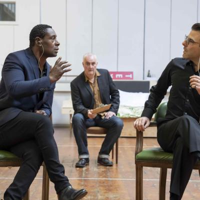 Actor David Harewood and Actor Zachary Quinto sit cross-legged looking at each other. Both actors wear earpieces and Harewood's hand is raised as if mid-speech. Actor John Hodgkinson sits in the background holding a clipboard while looking at Harewood. 