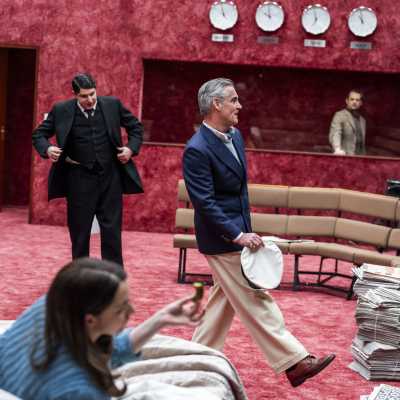 Four characters in frame. In focus there are two men. One in black suit adjusting his blazer, the other one in navy blazer holding a white cap in his hand they both seem happy. In the front of them a woman lying in bed in blue cardigan holding a pickle, blurred. Behind the two men, a men in beige blazer is standing behind the display window.	