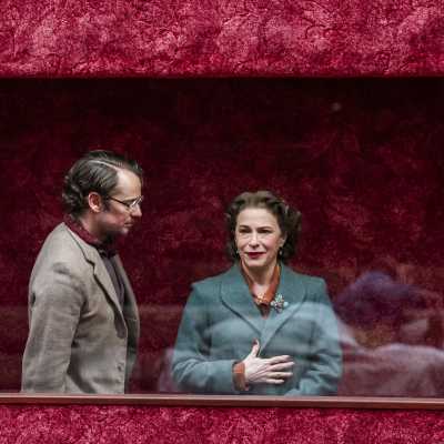 Two people standing behind a display window - a man with classes in grey blazer and a woman in teal coat. The woman is facing the front with her right hand on her upper abdoman and the man is looking at her facing side way 	