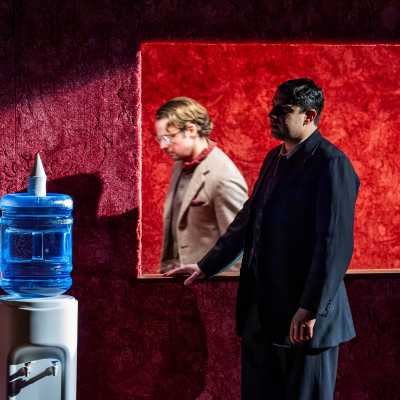 A man in black suit standing next to a water dispenser looking sideway. Another man in grey suit standing behind the display window, blurred.