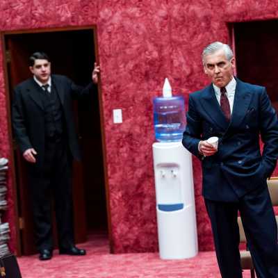 Two men in the photo, one in lighter colour suit standing in the focus with a cup in his hand, the other standing behind him by the door in black suit