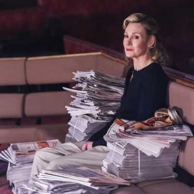 A woman with blonde hair wearing navy blue blouse sitting on a bench with piles of news papers around her	