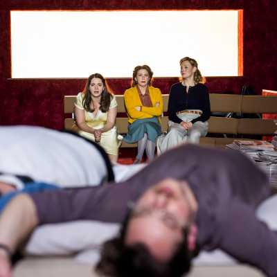 In focus, three women sitting on the bench, looking at two men lying in bed out, blurred	