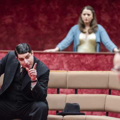 A man in three piece black suit sits on a bench looking and pointing  at the another man in white shirt	