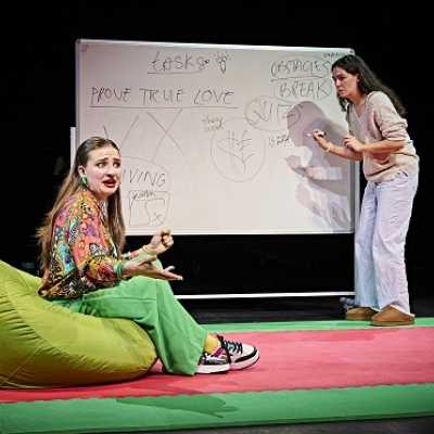 One person sits on a bean bag while another person stands writing on a whiteboard. 