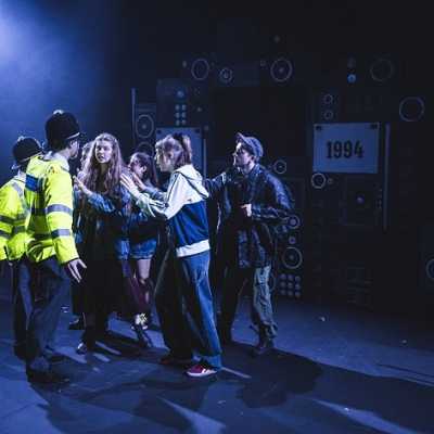 A group of people argue with two people dressed as police officers. Behind them a backdrop with images of speakers in black and white is visible.