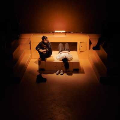 In a room lined with step-like blocks that jut out from the walls, a Caucasian man with sandy hair sits on a freestanding box on wheels with his knees pulled up around him. Next to him on the box lies a small, dark haired woman. Two pairs of shoes are on the ground next to them. The room is lit by one orange fluorescent bar that glows.
