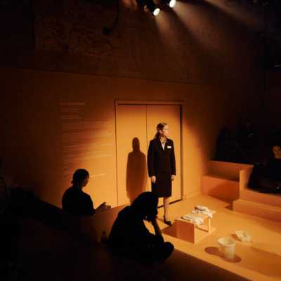 A white woman with tied-back blonde hair, wearing a dark suit, stands in front of a set of double doors under a spotlight. Around her, the room is lined with step-like blocks that jut out from the walls, some of which have people sitting on them. On one of the walls is some text like the kind that would be next to a painting in an art gallery.