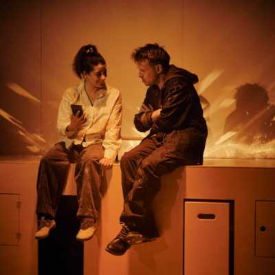 A small, dark haired woman with mid length hair in a ponytail and a Caucasian man with sandy hair and blue eyes sit on a cabinet built into an orange wall. They are both covered in custard. The woman holds up a small block-shaped object to show to the man while he speaks.