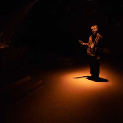 A Caucasian man with sandy hair and blue eyes holds a small block-shaped object. He is lit by an orange spotlight in an otherwise dark space.